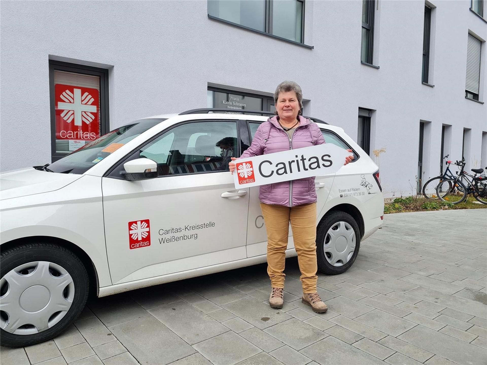 Ausfahrerin von Essen auf Rädern vor einem Caritas-Fahrzeug mit einem Caritas-Wegweiser in ihren Händen